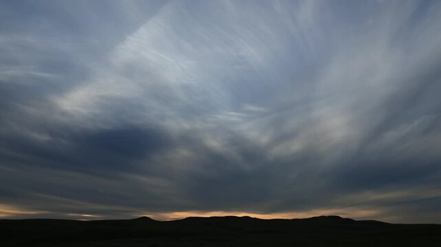 Timelapse of evening blue-gray clouds moving towards the viewer with a silhouetted horizon of rolling hills.
