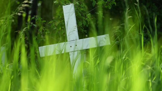 A weathered white wooden cross stands slightly tilted in a dense field of vibrant tall moving green grass and foliage.