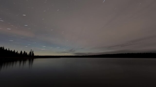 Night sky timelapse of clouds streaking across the sky and stars with tails rotating above a large calm northern lake

