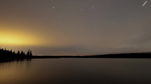 Timelapse of the night sky with stars and star-trails rotating above a large northern lake and clouds streaking across the sky
