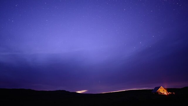 Time-lapse with tiny illuminated cabin rests under a vast, dark blue night sky filled with countless glowing stars and distant clouds.
