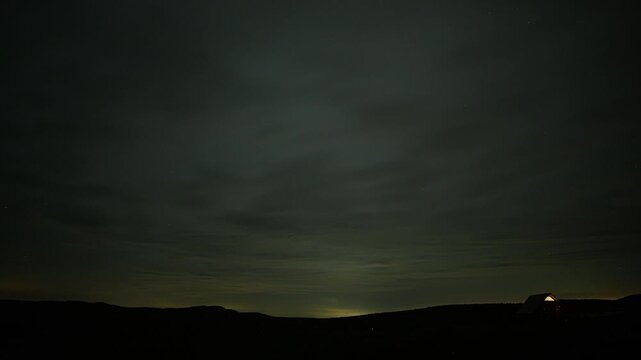 Timelapse Dark, heavy clouds roll over a tiny, glowing cabin under a deep purple and blue twilight sky.
