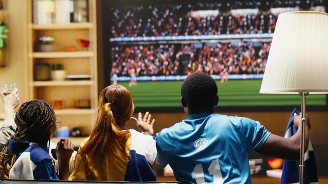 Football fans watch a match on the sofa with snacks and drinks ready for game night as high fidelity sports broadcast brings live stadium crowd energy into an apartment of friends.