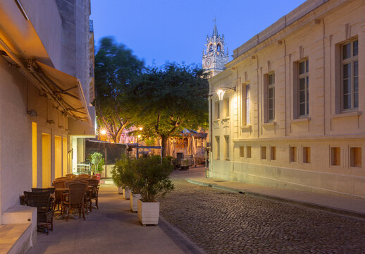 Dawn view of a quiet cobblestone street leading toward the clock tower in the historic center of Avignon, Provence, France.