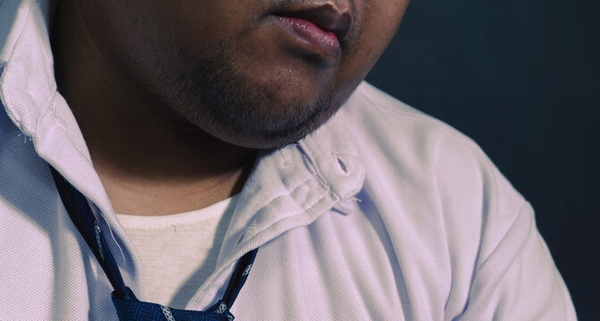 A moody close up captures the lower face and neck of an exhausted adult Asian male wearing a loose blue necktie and an unbuttoned white shirt. The wet sweating skin and rough stubble