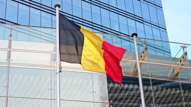 Belgian flag with black, yellow and red stripes waves beside a glass building, national symbol, european government building.