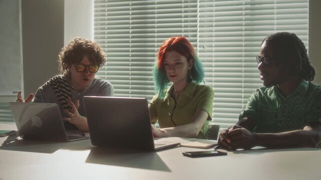 Diverse team of young creative professionals discuss business project details on laptop screen, share feedback and ideas during office meeting