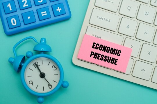 Pink sticky note with "ECONOMIC PRESSURE" text on a keyboard next to alarm clock and calculator. Conceptual metaphor for inflation, recession anxiety, budgeting under a tight deadline, or the general 