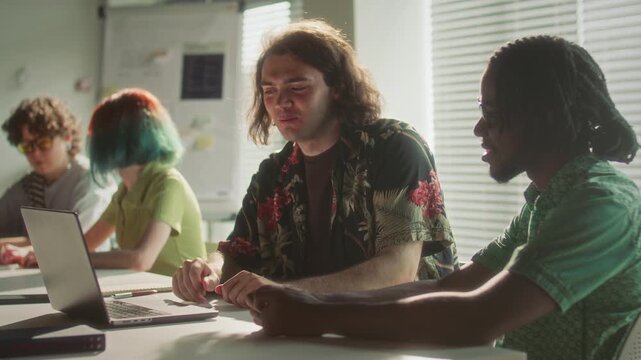 Young Caucasian man in floral shirt smiling and discussing data on laptop screen with his Black colleague during workday in sunlit open plan office