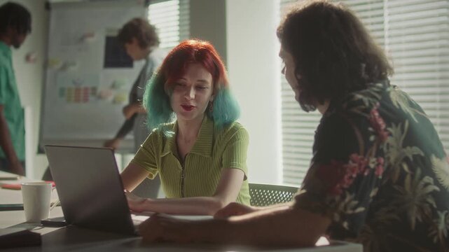 Creative gen Z businesswoman with colorful red and teal hair discussing data on laptop screen with her male colleague at desk in modern office, their coworkers talking by flipchart in background