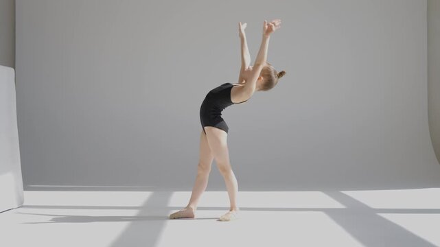 A little Caucasian girl gymnast in a black leotard does a backflip on a white background. 