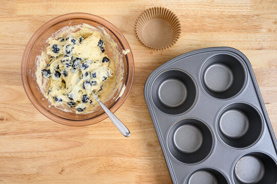 Muffin batter with fresh blueberries in a glass mixing bowl on wood counter, jumbo muffin tin with paper liners, baking lemon blueberry muffins at home
