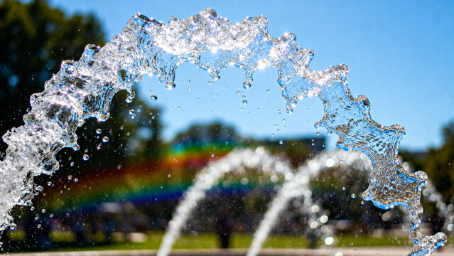 A fountain's water jet arching against a blue sky with sunlight creating glints.