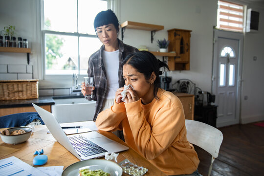 Sick woman working from home with laptop as partner offers water in kitchen