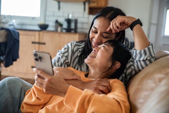 Two women laughing at a smartphone on a cozy sofa at home