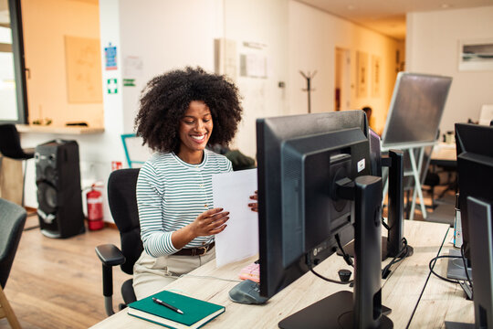 Smiling office worker reviewing documents at desktop computer in modern open-plan office