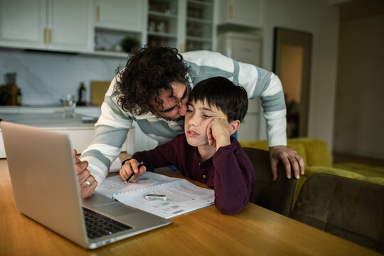 Father supporting bored son with homework at kitchen table