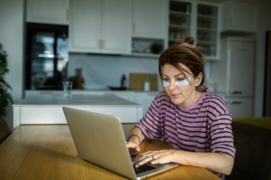 Young woman wearing eye patches working on laptop in home kitchen