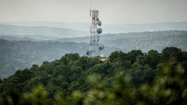 Focused medium shot of a microwave tower perched on a wooded hilltop surrounded by outoffocus dense forest and distant hills.