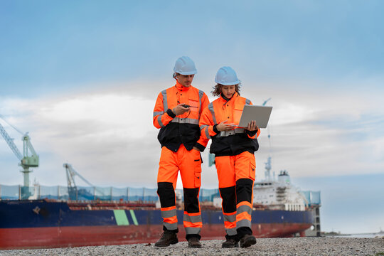 Professional maritime workers discussing operations with laptop at shipping terminal, Port engineers team in safety gear and high visibility vests walking together at dock