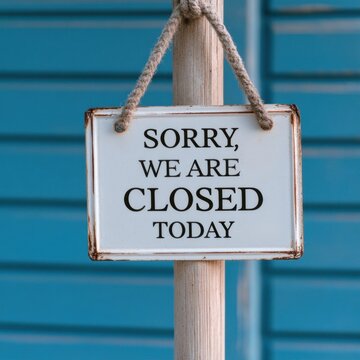 Rectangular white metal sign with black lettering reading 'sorry, we are closed today' hanging from a rustic wooden post against a blue wall