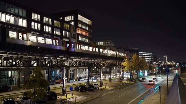 Subway train passing over elevated tracks above busy road intersection in city during dark night under bright street lights.