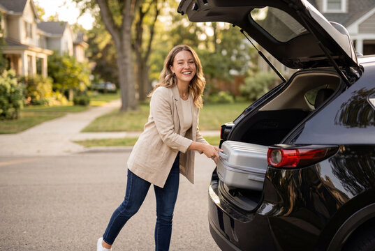 Happy young woman loading silver suitcase into black car trunk