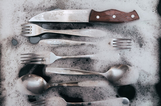 A silver knife, fork and spoons in the detergent foam on a black oven-tray. Washing dishes concept.