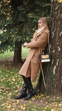 Vertical video. Nature relax. Autumn park. Dreamy woman in beige coat leaning against tree trunk closing eyes. Peaceful break outdoor with foliage on ground.
