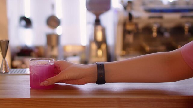 Drink order. Refreshing aperitif. Female customer hand reaching bright pink cold alcohol cocktail at pub counter in restaurant interior.