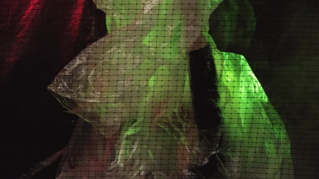 Stop danger. Ecology problem. Conceptual art. Plastic reuse. Woman putting off transparent polyethylene film stay free behind cell net red green background smoke blur copy space.