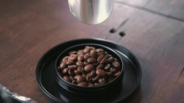 Roasted coffee beans falling from a metal scoop into a black container on a wooden table.