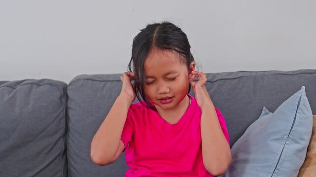 Little Asian girl covering her ears and grimacing from painful earache while sitting on sofa