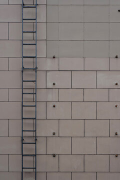 Building facade featuring beige rectangular cladding panels, exposed bolts, and a dark metal fire escape ladder.