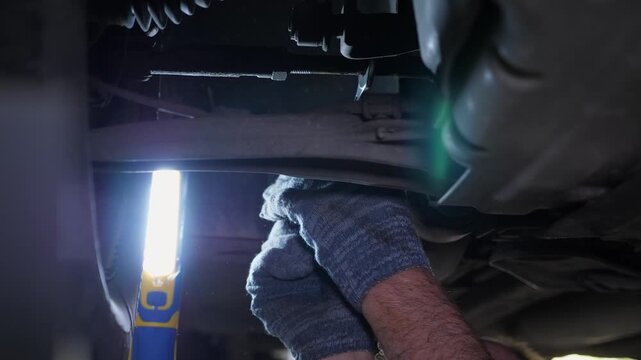 A detailed view of a mechanic working on a car, using a car lift in the garage. The mechanic is using a screwdriver to loosen the bolts on the underside of the car.