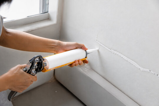 A person is applying caulk to a crack in a white wall indoors.