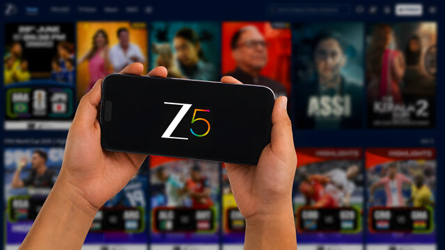 Hand holding smartphone showing Z5 OTT streaming app in front of laptop displaying Z5 website with Hindi content. Indian streaming platform editorial concept.
