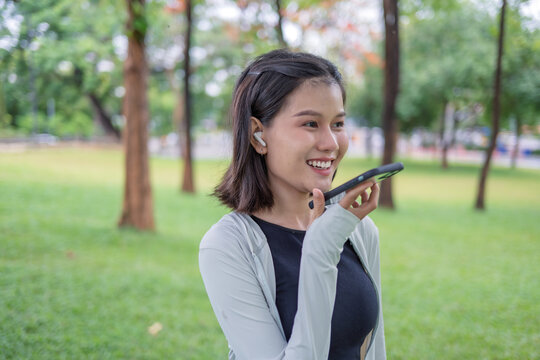Asian Gen Z woman using AI fitness tracking app on smartphone before jogging in green urban park.