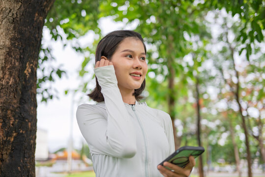 Asian woman with wireless earbuds warming up before jogging in green urban park for wellness lifestyle