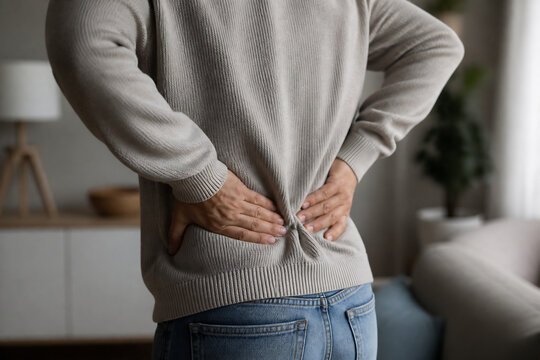 A person in a beige sweater holding their lower back while standing indoors.