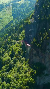 Vertical video. Altindere, Trabzon Province, Turkey. Sumela Monastery, a multi-storey stone complex with red-tiled roofs built directly into a sheer cliff face, aerial drone view over dense coniferou