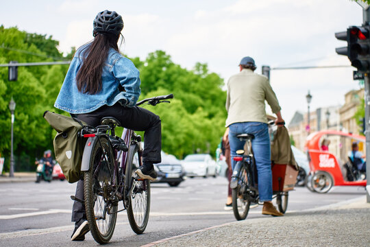People on bicycles waiting at a traffic light at a busy city intersection. Cyclists riding bike on city street with cars and green trees in the background