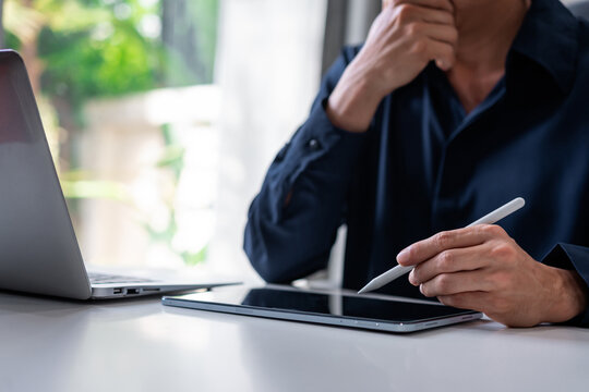 Thoughtful businessman holding a stylus pen over a digital tablet while working on a laptop at his desk