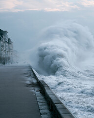 Fototapeta premium Massive wave crashes over sea wall with force. Sky clouds loom heavy, adding drama. City buildings line damp walkway. Faint figures walk cautiously near edge. Stormy sea power feels overwhelming