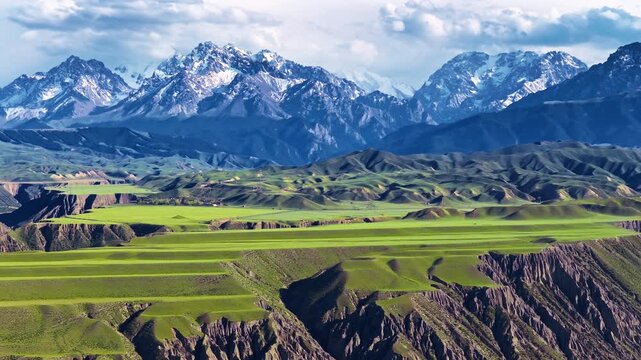 Vast green plateau with dramatic cliffs and snow capped mountains under a cloudy blue sky in Xinjiang, China.