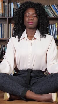 Calm african american businesswoman practicing yoga meditation in a library, sitting in a lotus position with eyes closed for stress relief, achieving mindfulness and well being at work