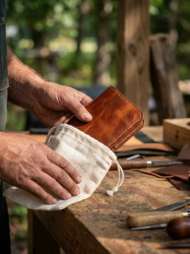 Leather craft hand placing handmade leather wallet into natural cotton bag on rustic wooden workbench with tools, artisanal packaging concept