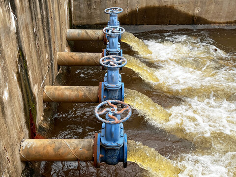 Industrial wastewater discharge facility close up, showcasing heavy duty blue control gate valves regulating brown effluent flow rate, hydraulic fluid pressure, and environmental drainage