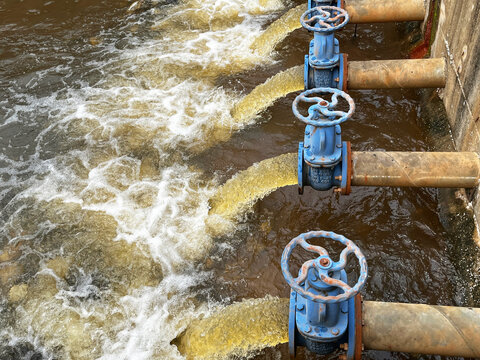 Industrial wastewater discharge facility close up, showcasing heavy duty blue control gate valves regulating brown effluent flow rate, hydraulic fluid pressure, and environmental drainage