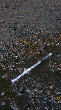 Heavy raindrops falling on a discarded hypodermic needle lying in a dirty puddle on wet ground, symbolizing social decay, drug abuse, and environmental pollution problems in urban areas